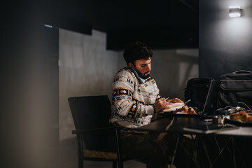 A young man concentrates on his writing in a dimly lit interior space, with a laptop and personal...