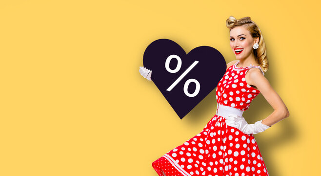 Woman holding black paper heart shape with % sign. Image of happy pin up girl. Blond model at retro fashion rockabilly rebates offers deals ad concept. Black Friday sale. Isolated yellow background.