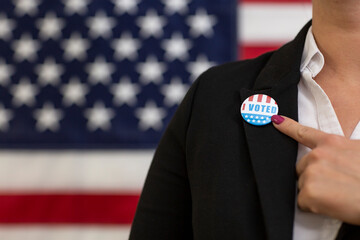 Young voter woman pointing with finger to i voted badge with USA flag in background. Political election concept