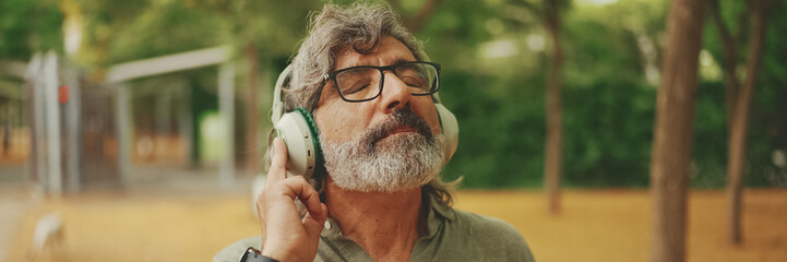 Friendly middle-aged man with gray hair and beard wearing casual clothes listening to music on headphones, Panorama. Mature gentleman in eyeglasses enjoys music outdoors