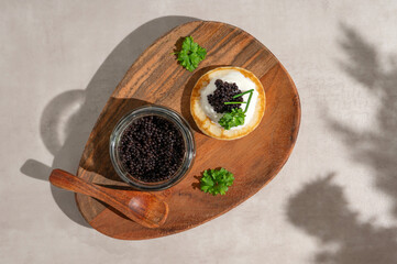 Black caviar on mini blini or pancake with glass jar on a wooden plate.