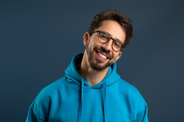 Smiling young man wearing glasses and bright hoodie standing against blue background