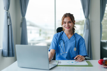 Friendly female nurse in blue scrubs seated at a desk, working on a laptop with medical charts and a stethoscope..
