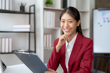 Young Asian professional business woman in a red suit reviews paperwork with a thoughtful expression in a well-organized office space..