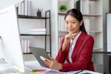 Professional young Asian businesswoman smiling while working on a digital tablet in a modern office setting.