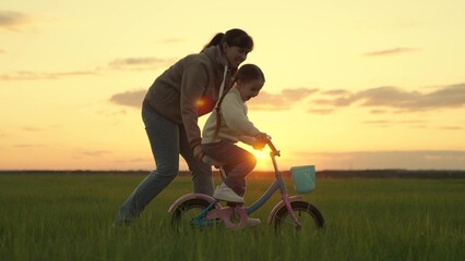 Mom teaches her little daughter to ride bicycle, sunset. Young mother teaches child to keep balance...