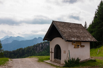 Fototapeta na wymiar Small chapel along scenic hiking trail on Monte Lussari in Camporosso, Friuli Venezia Giulia, Italy. Looking at majestic mountain ridges of Julian Alps. Wanderlust on pilgrimage route in European Alps