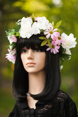 Naklejka premium Plastic woman mannequin with long black hair wearing a flower wreath and a black lace top outside in a blurry trees background