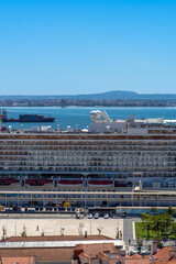 View of a large cruise ship docked in the port of Lisbon on the Tagus River with a container ship in the background.