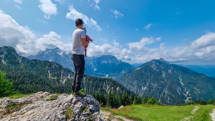 Naklejka premium Father holding baby with panoramic view from observation point of Monte Lussari, Camporosso, Friuli Venezia Giulia, Italy. Looking at mountain ridges of Julian Alps. Family trip on cloudy summer day