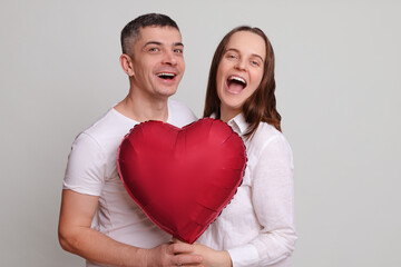 Fototapeta na wymiar Laughing man and woman wearing white clothing holding heart shaped air balloon isolated over gray background enjoying Valentine Day celebration expressing happiness