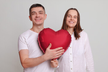 Pleased pretty couple looking at camera with smile satisfied man and woman wearing white clothing holding heart shaped air balloon isolated over gray background being in good mood