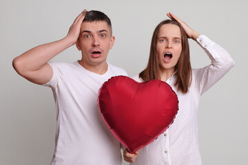 Fototapeta na wymiar Displeased confused man and woman wearing white clothing holding heart shaped air balloon showing facepalm gesture looking at camera with omg expression isolated over gray background