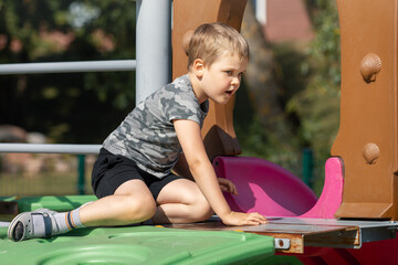 Fototapeta na wymiar A small boy in black shorts and a gray camouflage T-shirt climbs on the playground equipment