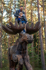 Fototapeta na wymiar A father and his son climb on a large elk sculpture in the park during a walk in the fresh air