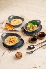 Three bowls of food are on a table, each with a different type of soup. The table is set with a fork, spoon, and knife, and there is a small ball on the table. Scene is casual and inviting