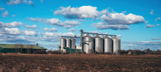 Agricultural silos of grain processing plant surrounded with plowed field, agriculture and farming buildings