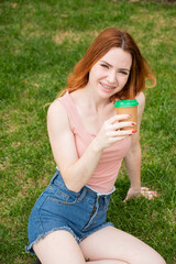 A beautiful young woman with braces on her teeth drinks from a cardboard cup while sitting on the grass. Vertical photo. 