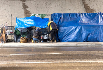 Homeless people at a camp with blue tarp and a car passing in the foreground during a rain storm in San Diego County Ca