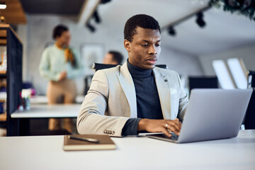 A black businessman sending some emails to his clients online, working at the office.