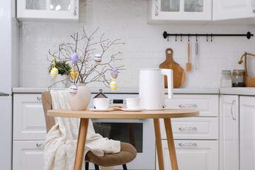 Home spring easter design in kitchen. White teapot, two cups of tea, plate with cookies, ceramic vase with branches and easter eggs on round wooden table, beige chair with cover in white kitchen.