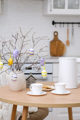 Home spring easter design in kitchen. White teapot, two cups of tea, plate with cookies, ceramic vase with branches and easter eggs on round wooden table, beige chair with cover in white kitchen.