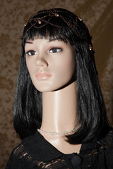 Plastic woman mannequin with black long hair wearing a black lace top and a chain head piece with beads posing on a golden brown background