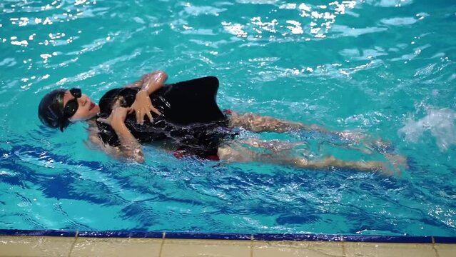 swimming pool training lesson for kids. little boy child learning to swim holding green floating board learning to move legs under water float on his side. child in swimming shorts, black cap and blue