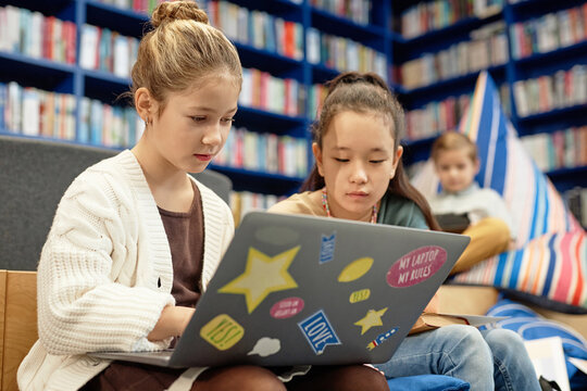 Portrait of two young girls using laptop computer in colorful school library and doing homework together