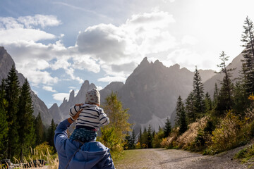 Mother carrying baby on shoulder along hiking trail on idyllic alpine meadow. Awe view of majestic mountain peaks of Sexten Dolomites, South Tyrol, Italy. Hike in panoramic Fischleintal, Italian Alps - obrazy, fototapety, plakaty