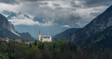 Panoramic view of moody landscape with small church in Dolomites mountains, Belluno, Veneto, Italy. Chiesa Parrocchiale di San Martino in Italian Alps in foggy and cloudy day at springtime