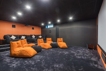 Luxury home theater with gray walls and carpet. Dark gray sofas and orange armchairs.