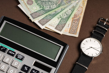 Polish banknotes PLN next to a lying calculator and watch with a white shield, brown background