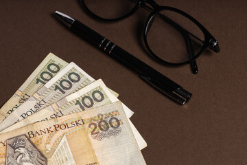 Polish banknotes PLN next to lying glasses and pen, brown background
