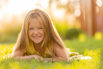 beautiful little girl with long blonde hair lies on a green lawn in the summer in a city park