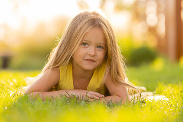 beautiful little girl with long blonde hair lies on a green lawn in the summer in a city park