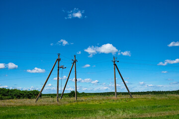 Three utility poles with wires against a blue sky with fluffy clouds. A green field and distant trees are visible.