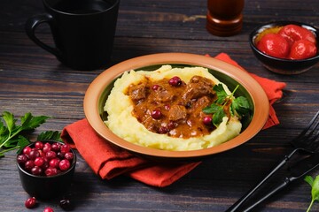 Roast beef stew with cranberries in sauce on a bed of mashed potatoes in a ceramic plate on a dark wooden background.