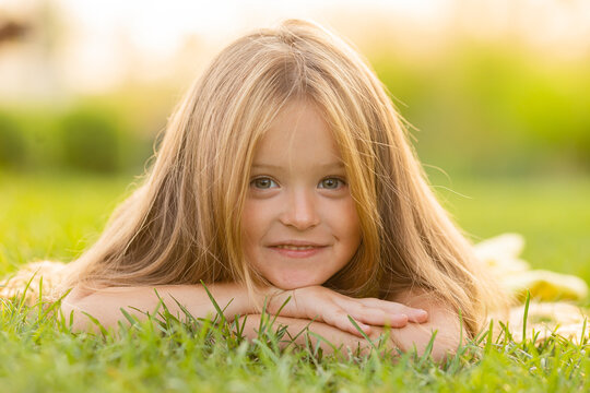 beautiful little girl with long blonde hair lies on a green lawn in the summer in a city park