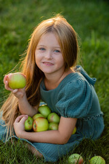 beautiful little girl with long blonde hair sits on the lawn in summer eating green apples