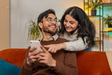 Smiling Arabian woman talking with boyfriend swiping smartphone sitting on sofa at home....