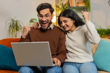 Cheerful young diverse couple in casual clothes using laptop surprised with email winning lottery...