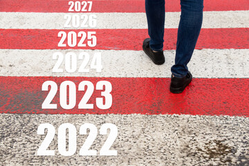 Male feet walk along a pedestrian crossing and on the transition of the year from 2022 to 2027 on...