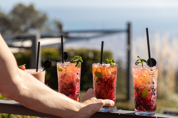 Hand picking up a fruit cocktail with a scenic ocean backdrop on a sunlit terrace - obrazy, fototapety, plakaty