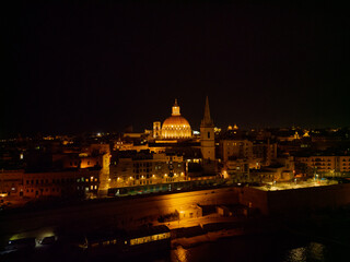 Fototapeta na wymiar Illuminated Basilica dome next to St. Paul's Cathedral at dark night. Aerial view from the sea towards Valletta old town. Residential buildings and streets with street lights.