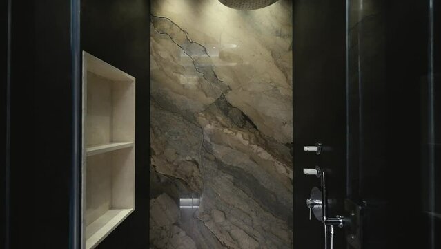 Elegant glass shower with amazing wall made of natural stone ceramic tiles equipped with white shelves and cozy lighting. Luxury plumbing and decorative elements in bathroom interior
