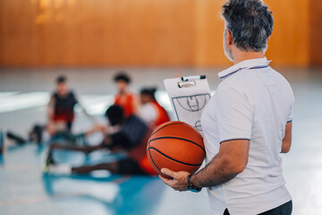 Rear view of a basketball coach with ball and clipboard in hands on court.