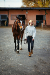 Young woman in civilian clothes leads her horse across the stables.