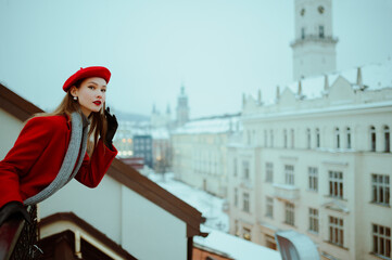 Fashionable woman wearing red beret, elegant winter coat, knitted gray scarf, gloves, posing on balcony with beautiful view on snow covered European city Lviv. Copy, empty, blank space for text