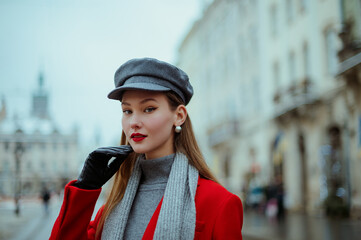 Fototapeta premium Fashionable confident woman wearing elegant red coat, gray cap, silver earrings, black leather gloves, posing in street of European city. Outdoor close up portrait. Copy, empty, blank space for text 